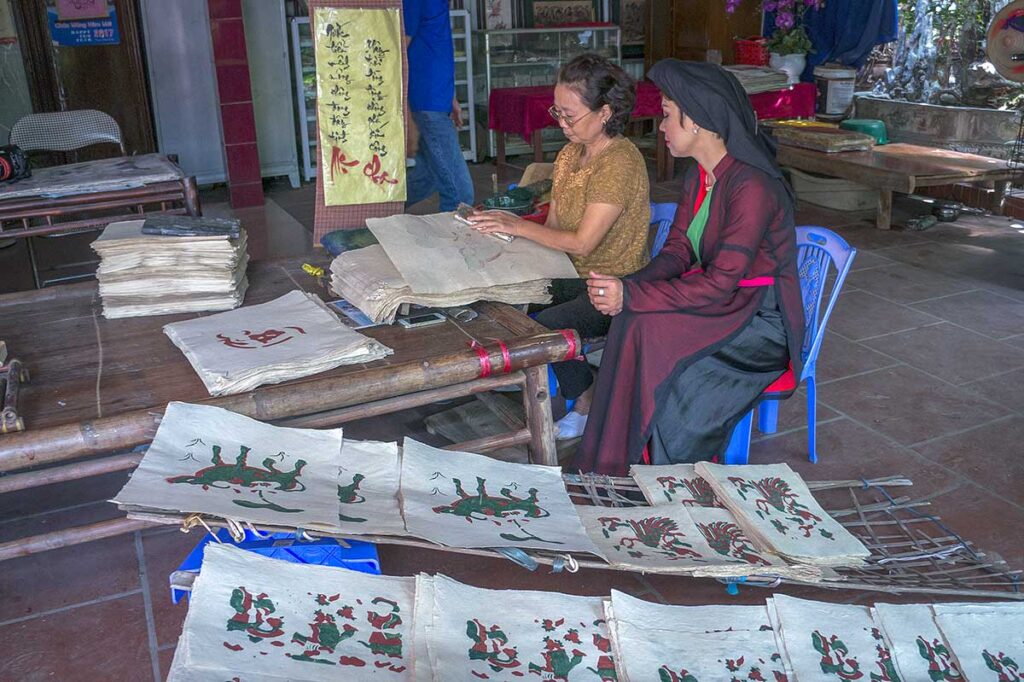 In the foreground a few Dong Ho Painting and in the background two woman making them at Kids are sitting on a mat with a man in traditional Vietnamese clothes and showing the children how to make a traditional Dong Ho Painting  during a workshop in Dong Ho Painting Village