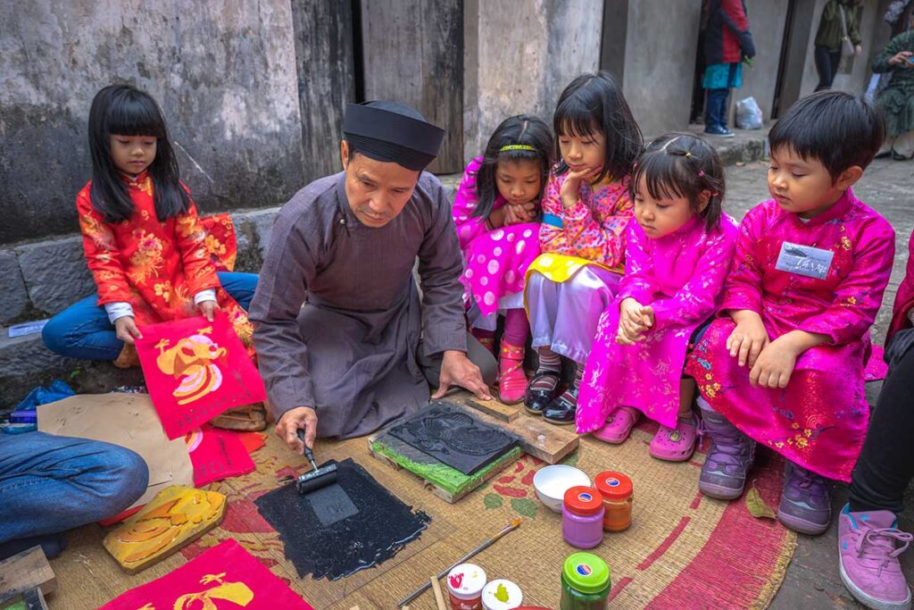 Kids are sitting on a mat with a man in traditional Vietnamese clothes and showing the children how to make a traditional Dong Ho Painting during a workshop in Dong Ho Painting Village