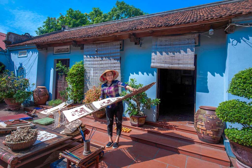 A woman standing in front of a local house and holden a bamboo drying rack with Dong Ho paintings at Kids are sitting on a mat with a man in traditional Vietnamese clothes and showing the children how to make a traditional Dong Ho Painting  during a workshop in Dong Ho Painting Village