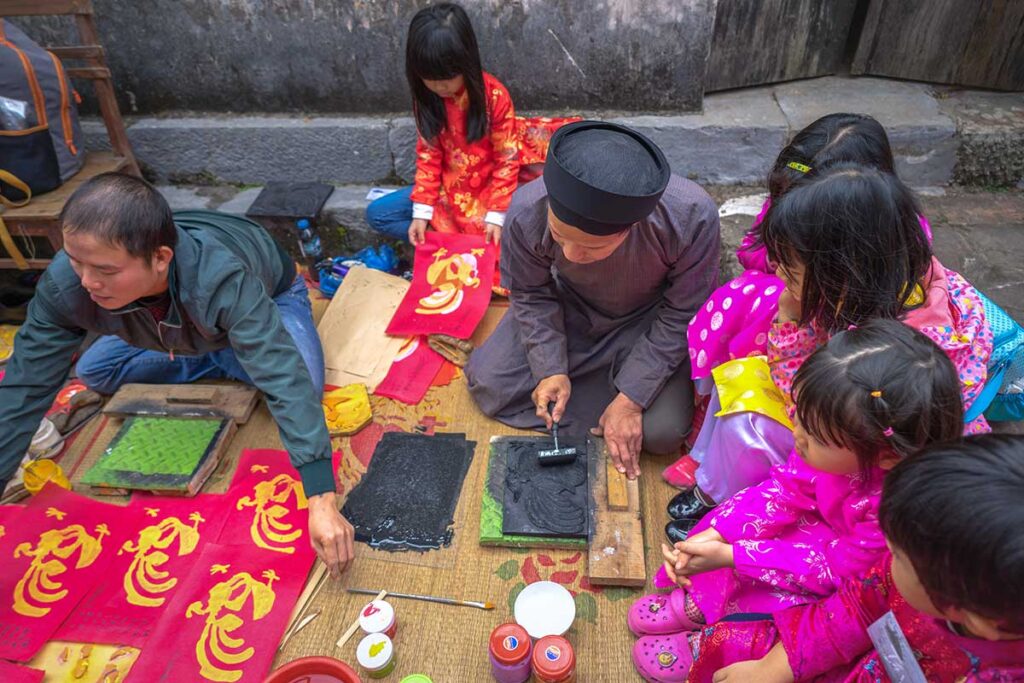 Kids are sitting on a mat with a man in traditional Vietnamese clothes and showing the children how to make a traditional Dong Ho Painting  during a workshop in Dong Ho Painting Village