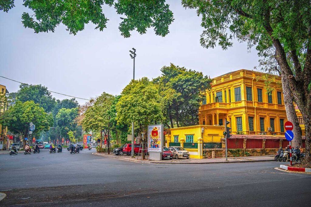 A wide street with trees and a yellow French colonial building in the French Quarter in Hanoi