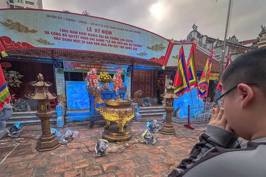 A man praying at an outside altar decorated with flags and a banner during  Hai Ba Trung Temple Festival