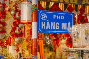 A street sign saying Pho Hang Ma with on the background all kind of joss paper and festival items for sale at a local shop