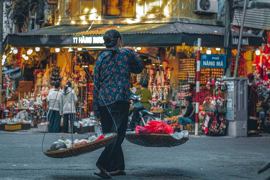 A Vietnamese street vendor carrying a shoulder pole with baskets of food at Hang Ma Street in the Old Quarter
