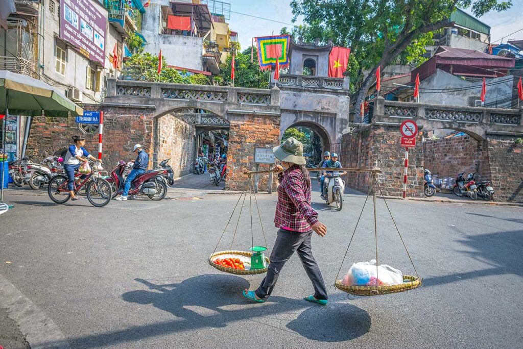A woman with stick on her shoulders carrying baskets of food walking in front of the Old City Gate of Hanoi