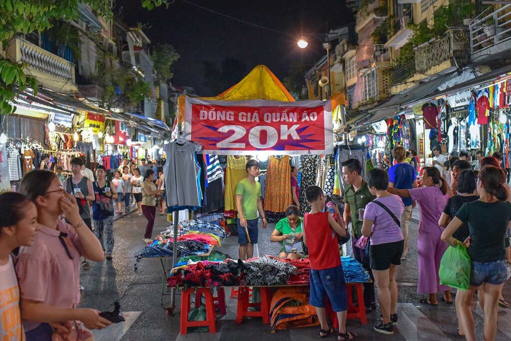 Lots of people walking between the stalls of Hanoi Weekend Night Market with a big Sale sign at one of the stalls with 20k Vietnam Dong 