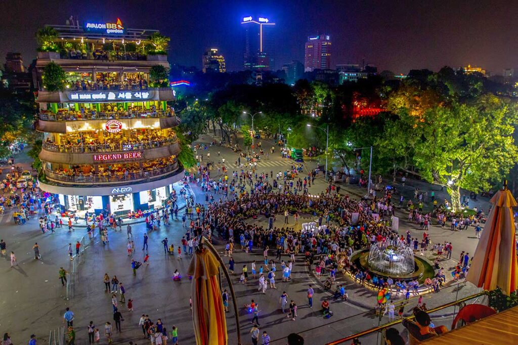 Seen from above, a large group of people in a circle looking at a local performance nearby Hoan Kiem Lake during Hanoi Weekend Night Market