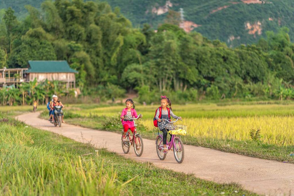 Local kids are cycling over a countryside line with Mai Chau rice fields on both sides near Lac Village