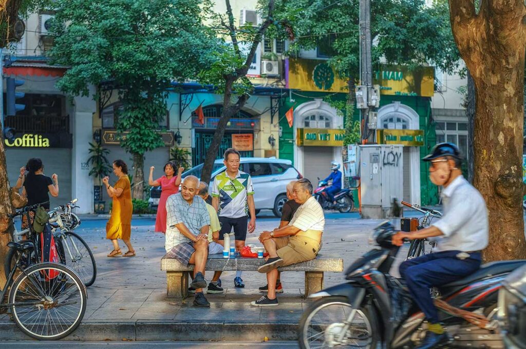 Local people of Hanoi - Vietnamese man sitting on a bench watching traffic and motorbikes coming by while woman on the background doing tai chi