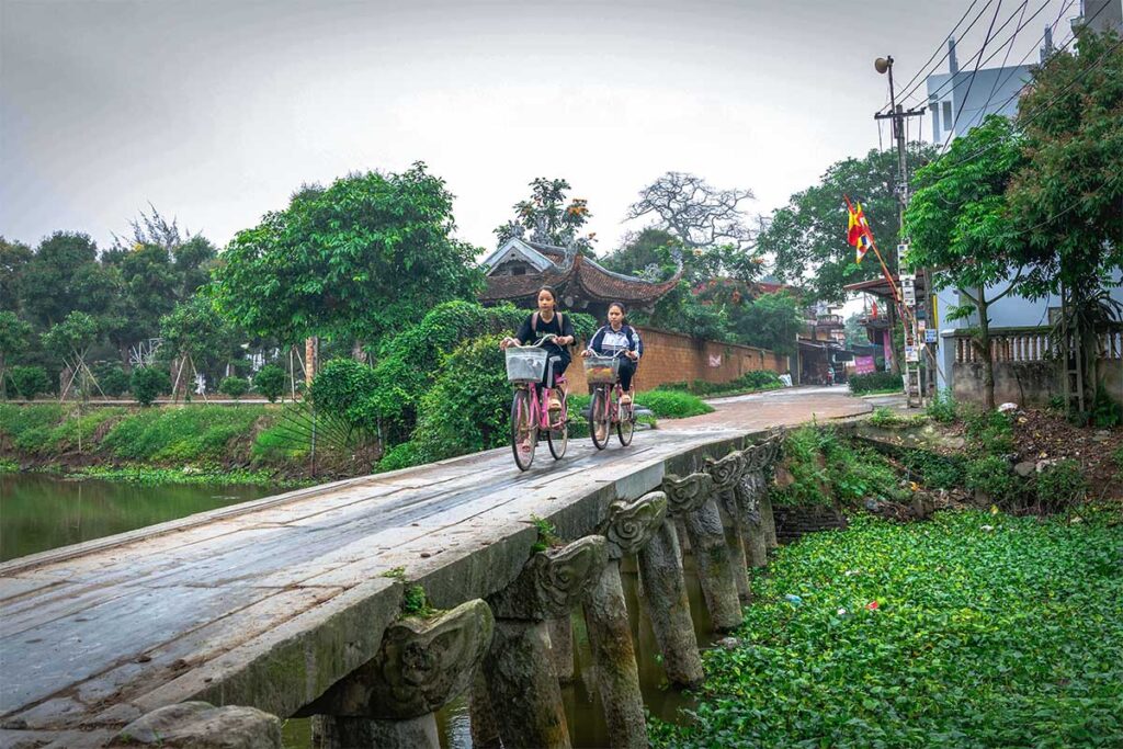 Two girls cycling over a stone bridge in Nom Village, Hung Yen