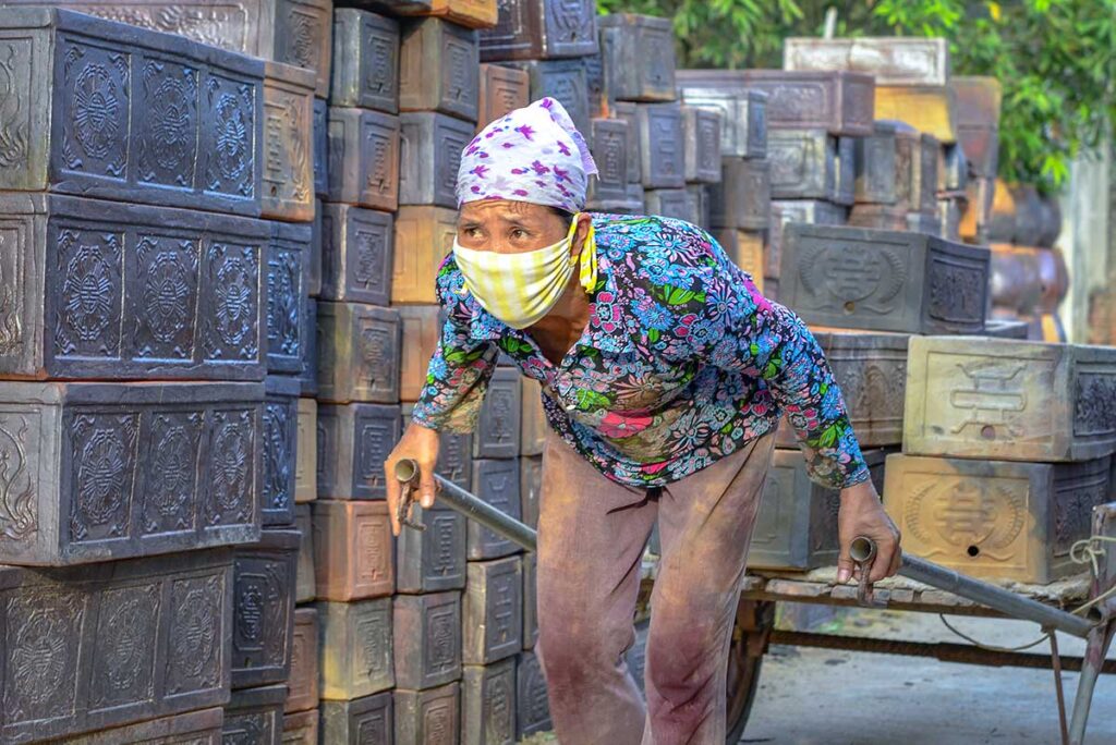 A woman with mask is pulling a cart with on the background clay square pots stacked at Phu Lang Potter Village