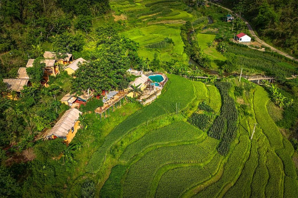 An aerial view of Pu Luong Riverside Lodge with bungalows and a pool overlooking the terraced rice fields of Pu Luong
