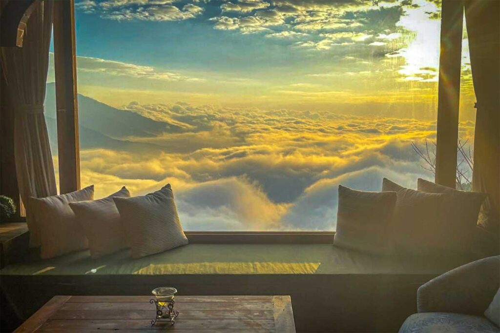 A large open window of a homestay in Ta Xua with pillows in the windowsill with outside views of the sunrise and hanging clouds below