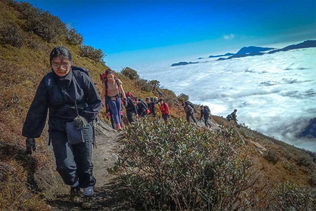 A group of tourists are trekking in Ta Xua Nature Reserve over a mountain path with in the background below them a sea of clouds