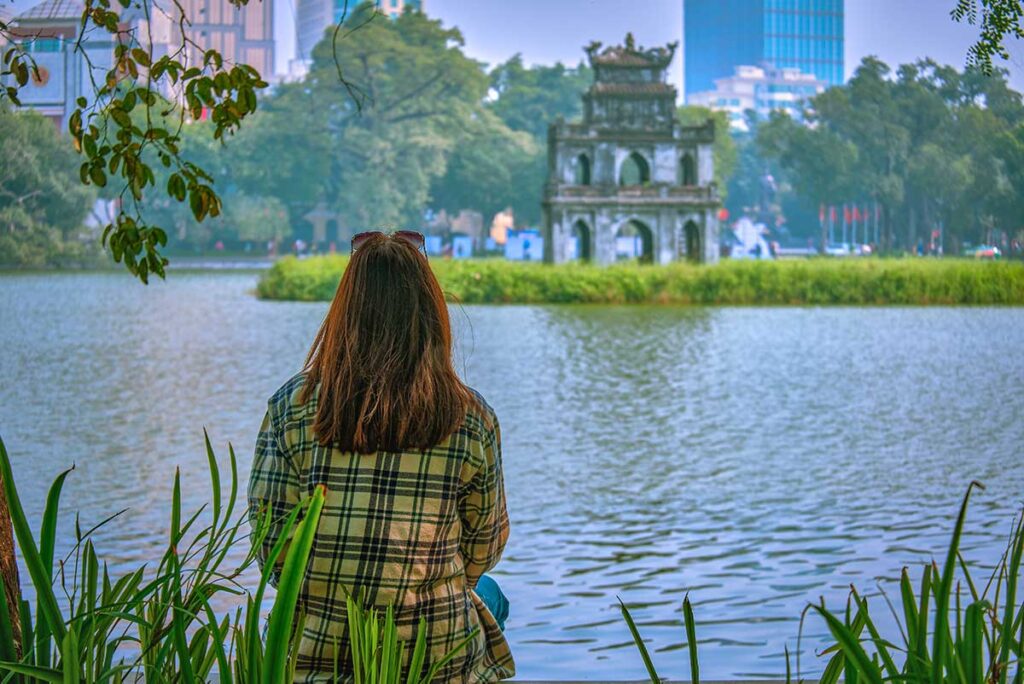 A girl sitting along the edge of Hoan Kiem Lake with on the background Turtle Tower on an island in the lake