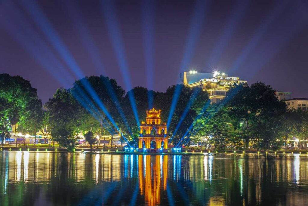 The Turtle Tower in Hoan Kiem Lake in the late evening when dark, with soft yellow lights illuminating the building and light beams from the island shining in the air