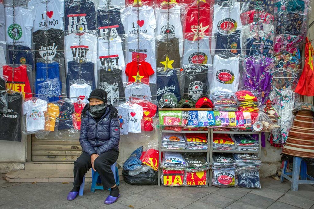 A woman in winter jacket sitting on the street of Hanoi selling Vietnam themed t-shirts to tourists