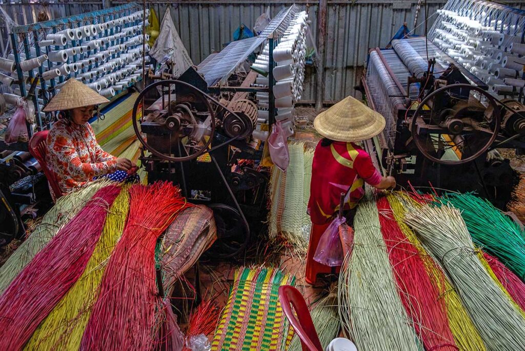 Two women weaving multicolored sedge mats on looms inside a workshop at Dinh Yen Mat Weaving Village