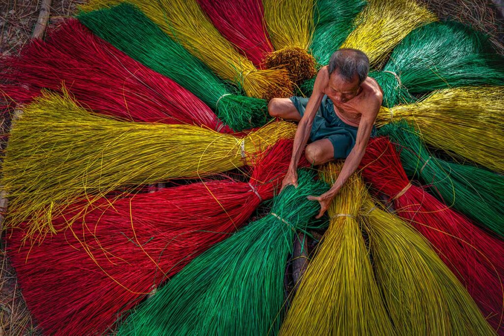 Elderly craftsman arranging bundles of dyed sedge grass into circular patterns at Dinh Yen Mat Weaving Village