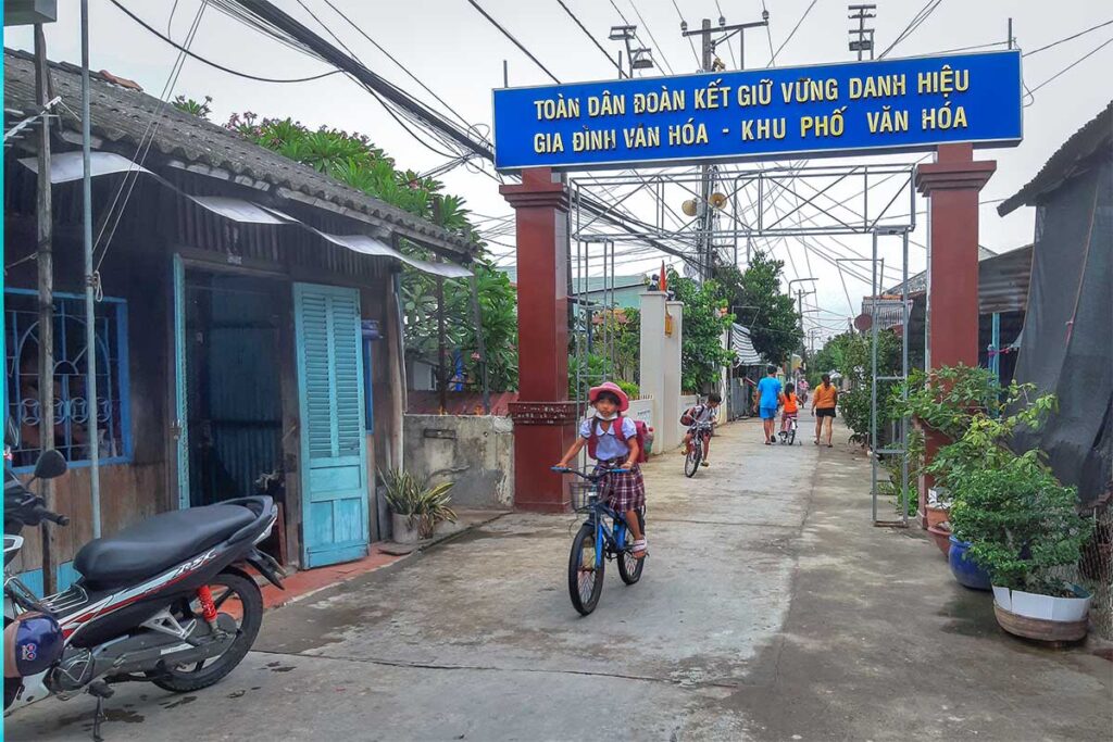 Village street with children cycling under a community gate on Dragon Island (Tân Long), My Tho City.