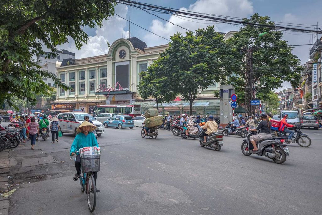 Hang Da Market seen from outside and the street filled with motorbikes, a few cars and a local woman biking through the street
