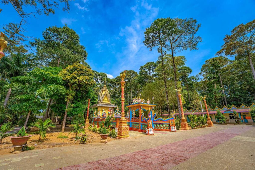 Courtyard of Hang Pagoda with colorful shrines, Khmer statues, and tall forest trees in the background.