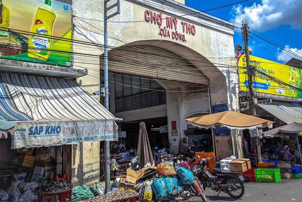 Entrance to My Tho Central Market in Tien Giang with vendors and motorbikes outside.