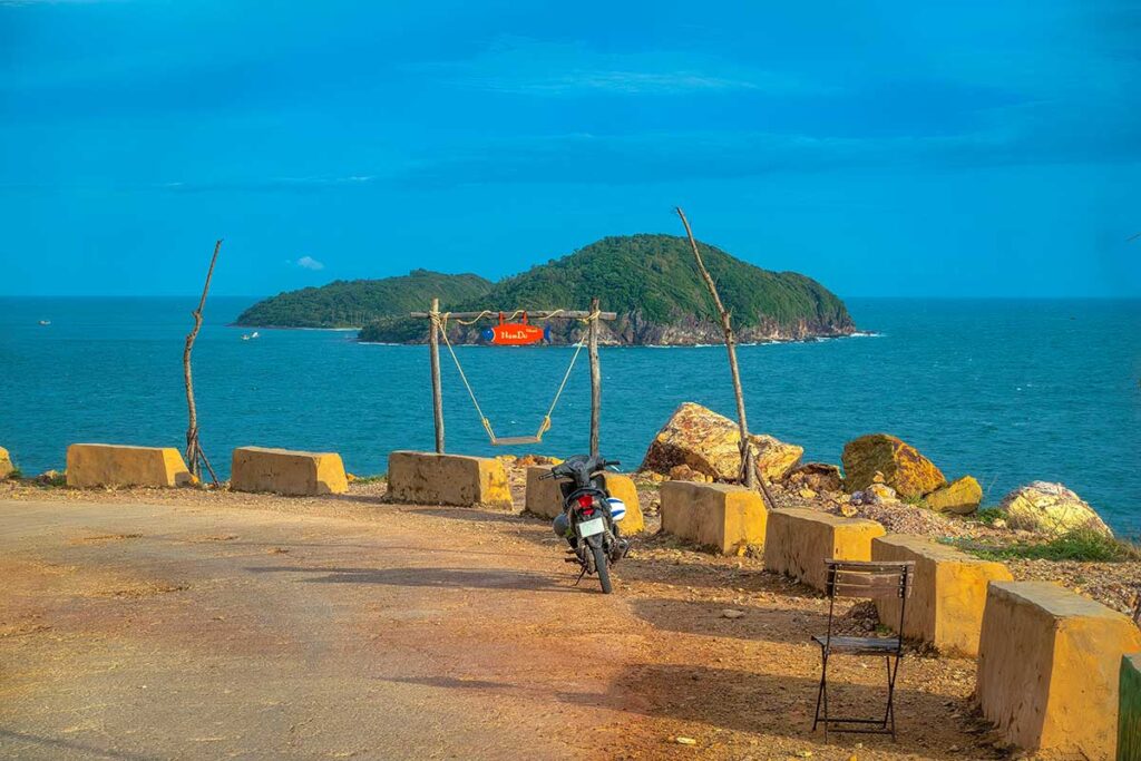 Scenic coastal road on Nam Du Island – Motorbike stop at a rustic swing facing Hon Dau islet, with deep blue ocean views along the Nam Du coastal loop.