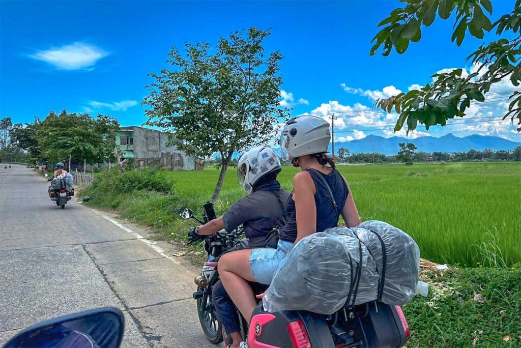 A foreign girl traveller sitting on the back of a motorbike of an easy rider that travels through the countryside with rice fields in the background from Phong Nha to Hue