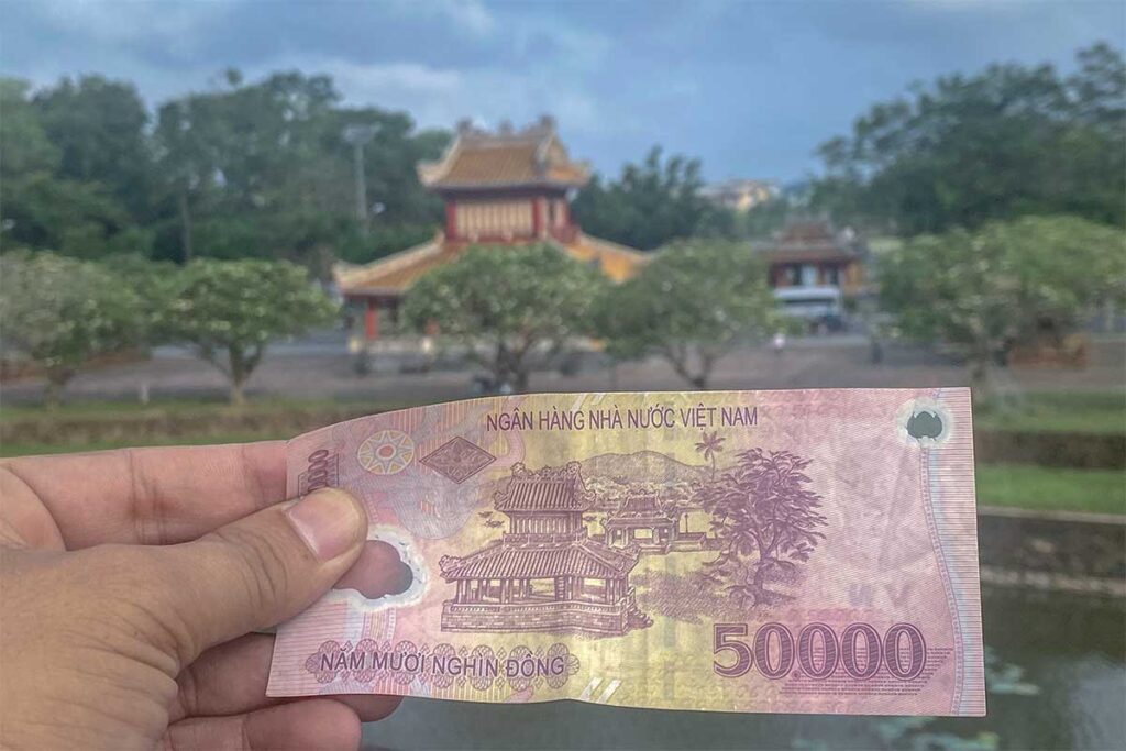 A hand holden a 50000 Vietnam Dong banknote in the background you see Phu Van Lau Pavilion and even further blurry Nghênh Lương Pavilion