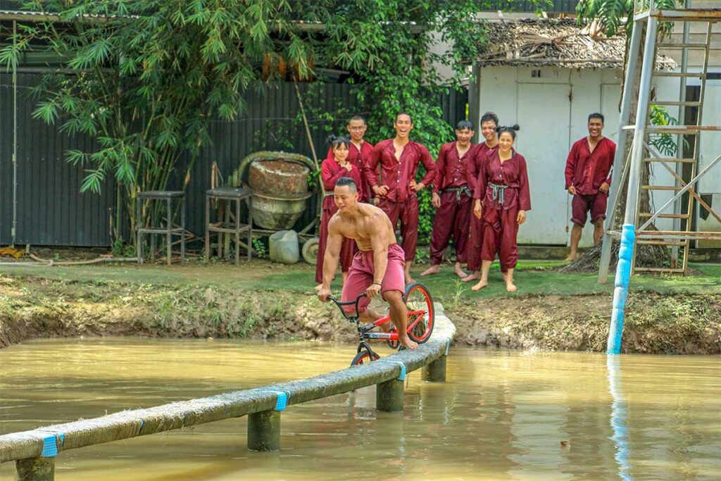 Tourists trying a folk game by cycling across a narrow bridge over a pond on Phoenix Island, Mekong Delta.