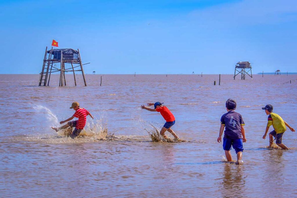 Children playing in the shallow muddy waters of Tan Thanh Beach Tien Giang, with stilted fishing huts in the background.