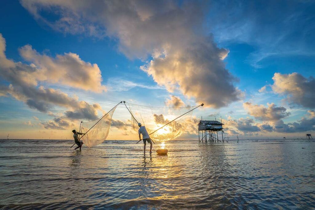 Fishermen lifting large nets at sunrise on Tan Thanh Beach Tien Giang, with a wooden stilt hut in the background.
