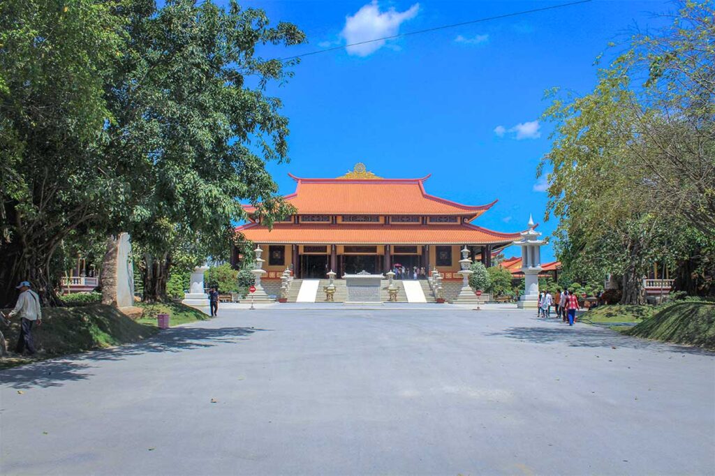 Main prayer hall of Trúc Lâm Chánh Giác Monastery in Tiền Giang with red-tiled roof and broad courtyard, framed by shady trees.