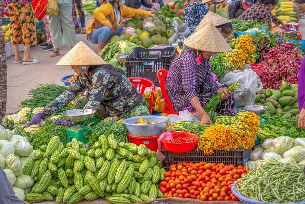 Female vendors at Vi Thanh Cho Hom arranging fresh herbs, flowers, tomatoes, and gourds on the ground