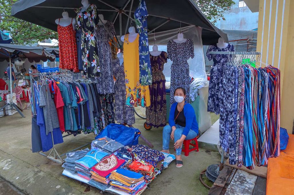 Clothing vendor at Ba Le Market in Hoi An displaying colorful dresses, T-shirts, and fabrics at an outdoor stall.