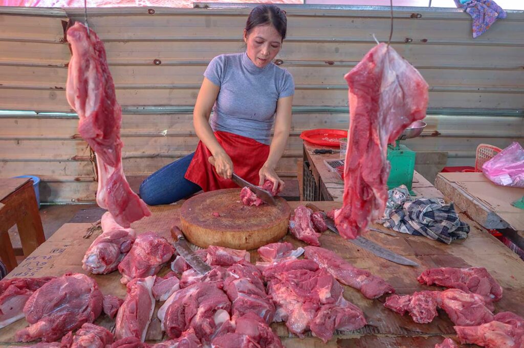 Local butcher cutting fresh pork on a wooden block at Ba Le Market in Hoi An, with hanging meat cuts and a traditional market stall setup.
