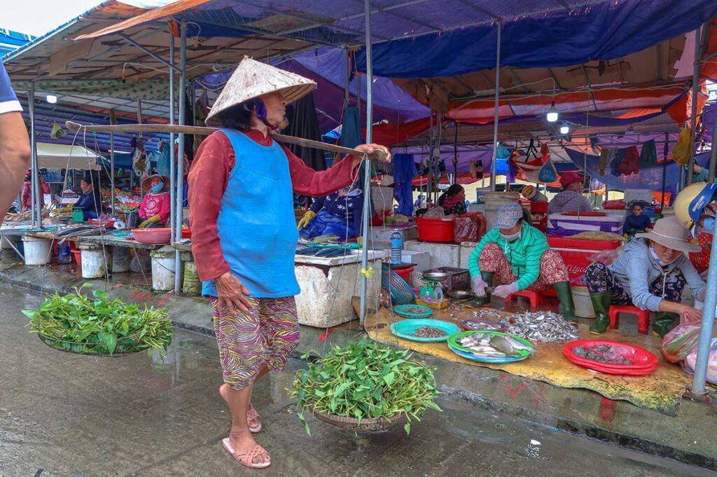 Elderly woman with a bamboo shoulder pole carrying fresh morning glory vegetables at Ba Le Market in Hoi An, passing the seafood section.