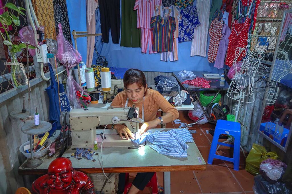Seamstress working on a sewing machine at Ba Le Market in Hoi An, offering tailoring and clothing repair services among colorful fabrics.