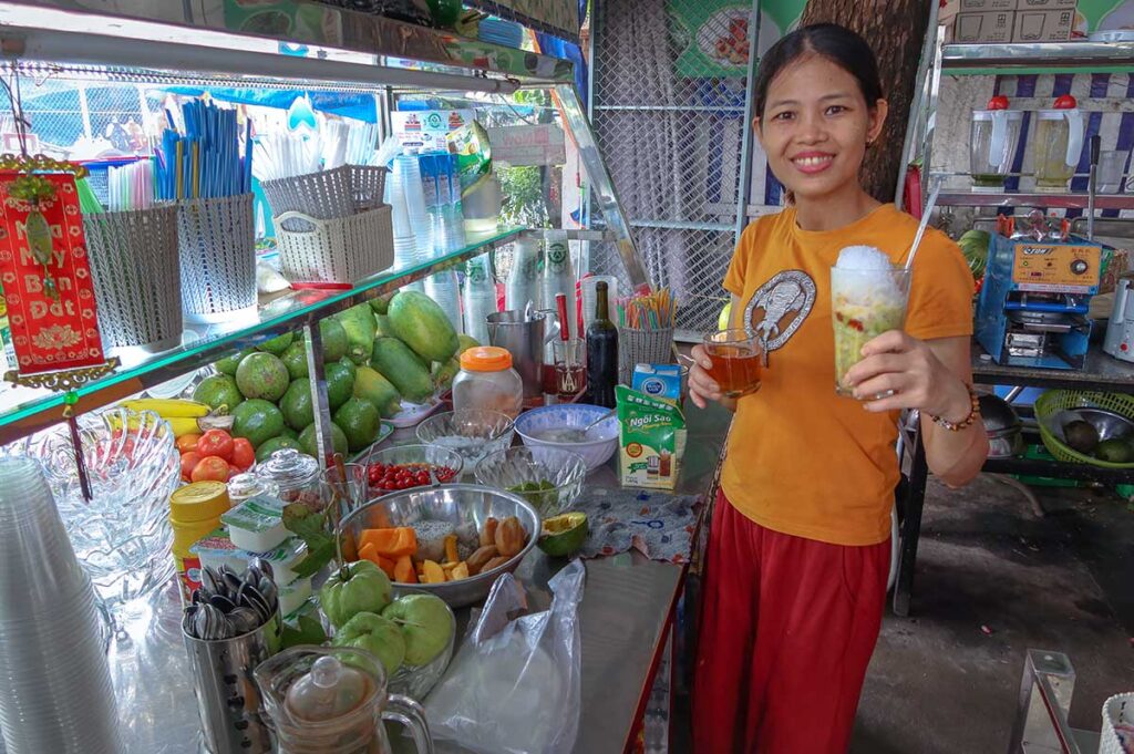 Smiling vendor at Ba Le Market in Hoi An serving a glass of Vietnamese chè dessert with crushed ice, surrounded by fresh tropical fruits and ingredients.