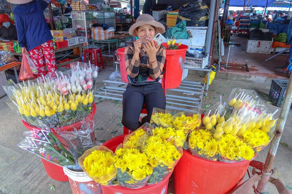 Flower seller at Ba Le Market in Hoi An with bright yellow chrysanthemums and lotus buds, commonly used for offerings and local celebrations.