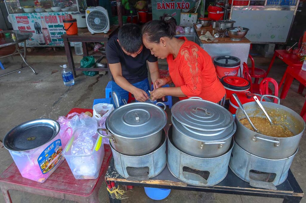 Street food vendor at Ba Le Market in Hoi An preparing local dishes from large pots, serving meals to customers at simple tables.