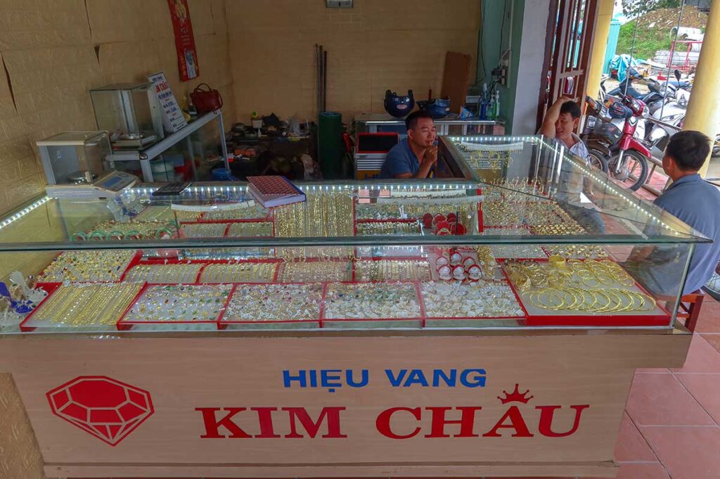Gold jewelry stall at Ba Le Market in Hoi An, displaying bracelets, rings, and necklaces under glass counters.