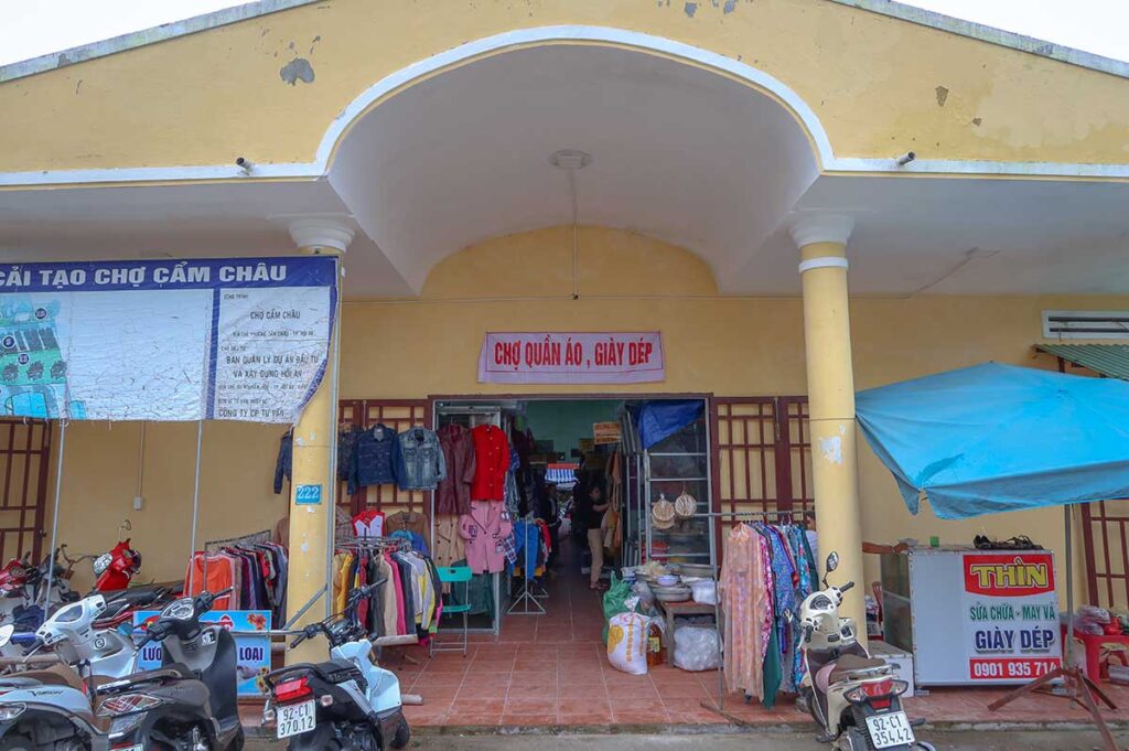 Front entrance of Ba Le Market in Cam Chau ward, Hoi An, with clothing stalls, scooters parked outside, and a yellow-painted market building.
