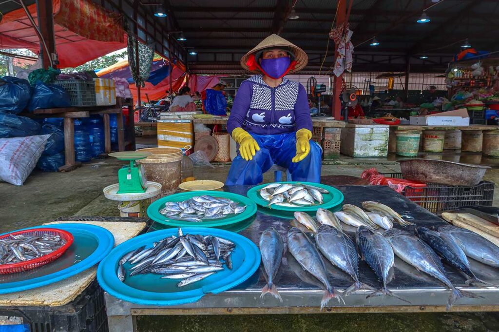 Fishmonger at Ba Le Market in Hoi An selling fresh tuna and small fish on metal trays, wearing a conical hat and gloves.