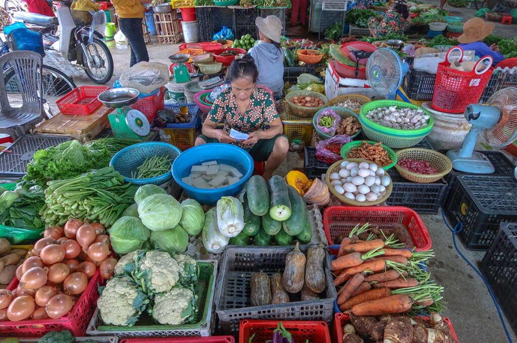 Vendor at Ba Le Market in Hoi An selling cabbage, carrots, tofu, eggs, and leafy greens, showcasing the diversity of local vegetables.