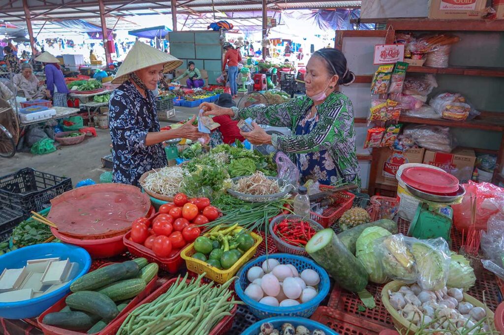 Woman buying fresh vegetables from a vendor at Ba Le Market in Hoi An, surrounded by tomatoes, cucumbers, herbs, and daily produce.