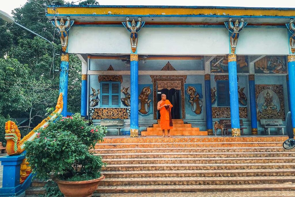 Monk in orange robe standing at the sala building of Bat Pagoda (Chùa Dơi) in Soc Trang, with blue columns and Khmer carvings.