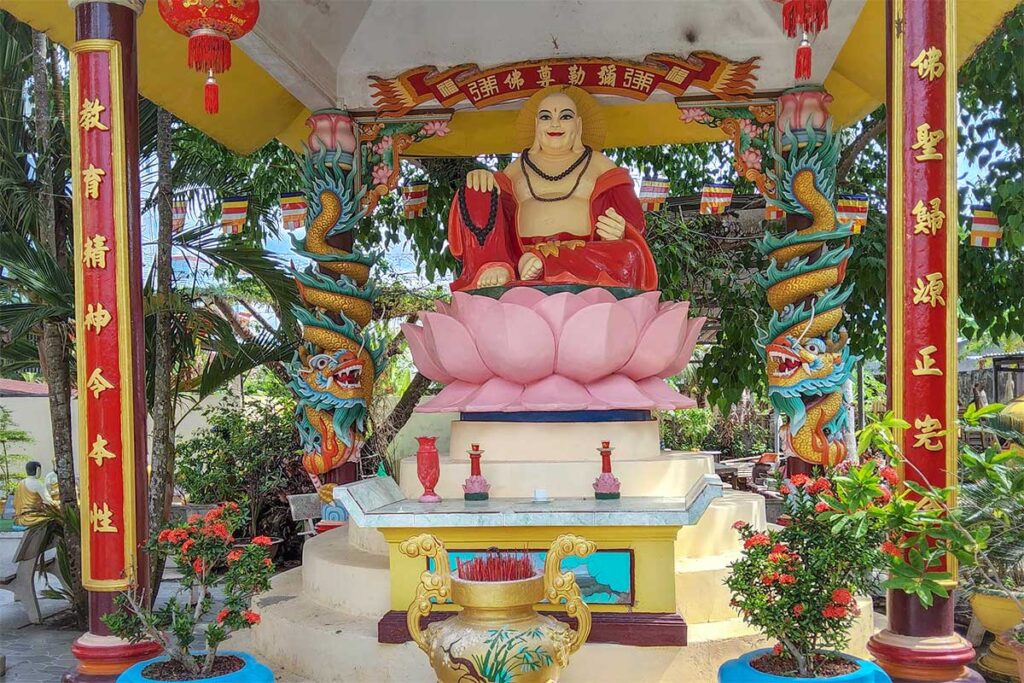 Outdoor Maitreya Buddha statue at Clay Pagoda – Large clay Maitreya seated on a pink lotus pedestal under a shrine roof decorated with red columns and dragons.