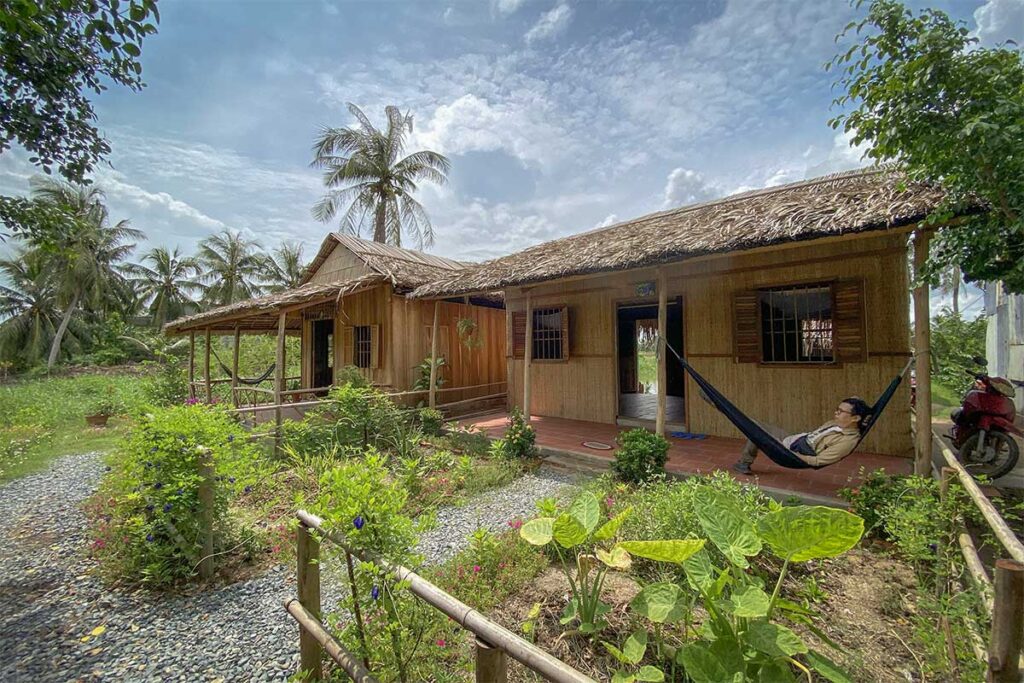 Rustic homestay with thatched roof and hammock in the garden on Con Chim Island, Tra Vinh, surrounded by coconut trees and flowers